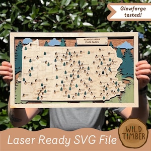 May include: A layered wooden map of Pennsylvania State Parks featuring engraved locations and small tree icons. The map includes a rectangular sign labeled Pennsylvania State Parks, blue clouds, and dark green forest silhouettes in the background.