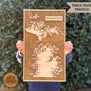 May include: Wooden map of Michigan with lighthouse markers. The map is framed in a dark wood and features the text "Michigan Lighthouses" and "Track Your Travels!". The Wild Timber logo is in the lower left corner.