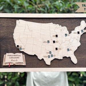 May include: Wooden map of the United States with a light wood frame. The map features a dark brown background and a lighter wood cutout of the US. Small black and blue squares mark locations. Text on the map reads "Track Your Travels!" and "Presidential Libraries Bucket List."
