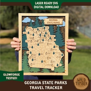 May include: Wooden Georgia State Parks travel tracker map. The map is laser-cut with a light wood finish, featuring a detailed outline of Georgia with labeled parks and green tree accents. The frame is light wood, with text that reads "Georgia State Parks Travel Tracker".