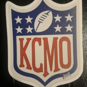 Può includere: Un adesivo a forma di scudo con la scritta "KCMO" in rosso. La parte superiore è blu con un pallone da football bianco e sette stelle bianche. L'adesivo ha un bordo bianco. Il design ricorda un logo sportivo.
