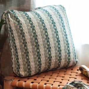 May include: A green and white patterned throw pillow with a woven texture. The pillow is sitting on a woven wicker chair.