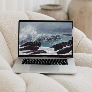 May include: A silver MacBook Pro laptop displaying a seascape image with crashing waves and dark rocks. The laptop is open and resting on a cream-colored, textured armchair. The screen shows the text "SIMPLYALLO".