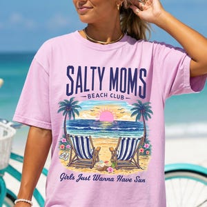 May include: A light pink short-sleeve t-shirt featuring a graphic of two beach chairs on sand facing the ocean at sunset. The text reads SALTY MOMS BEACH CLUB and Girls Just Wanna Have Sun. The design includes palm trees and tropical flowers.