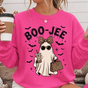 May include: A bright pink crewneck sweatshirt featuring a graphic of a ghost wearing leopard print sunglasses and a matching bow. The ghost holds an iced coffee and a leopard print handbag. The text BOO-JEE is printed above with black bats.