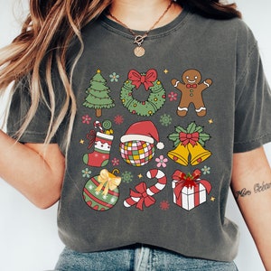 May include: A dark gray t-shirt with a Christmas theme. The shirt features a variety of festive icons, including a Christmas tree, a wreath, a gingerbread man, a stocking, a candy cane, a present, a bell, and a Christmas ornament. The text "Merry & Bright" is printed on the shirt.