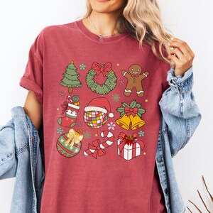 May include: A crimson short-sleeve t-shirt featuring a grid of colourful Christmas-themed illustrations including a tree, wreath, gingerbread person, stocking, disco ball with a Santa hat, bells, ornament, candy cane, and a gift box.