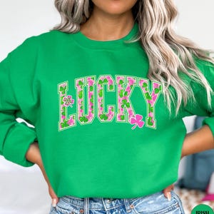 May include: A green sweatshirt featuring the word "LUCKY" in a white outline, filled with a pink floral and shamrock design. A pink shamrock is on the right. The sweatshirt is a solid green colour.