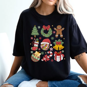 May include: A black short-sleeved t-shirt featuring a colourful graphic print of various Christmas icons including a tree, wreath, gingerbread person, stocking, disco ball with a Santa hat, bells, ornament, candy cane, and a gift box.