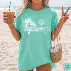 May include: A mint green short-sleeved t-shirt featuring a white line art graphic of palm trees, a sun, and ocean waves. The shirt has a crew neckline and a relaxed fit. The graphic is positioned centrally on the chest area of the garment.