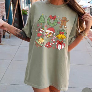 May include: A khaki crew neck t-shirt featuring a graphic print of various Christmas icons including a tree, wreath, gingerbread person, stocking, disco ball with a Santa hat, bells, ornament, candy cane, and a gift box with a red bow.