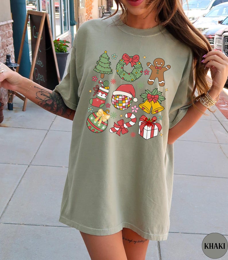 May include: A khaki crew neck t-shirt featuring a graphic print of various Christmas icons including a tree, wreath, gingerbread person, stocking, disco ball with a Santa hat, bells, ornament, candy cane, and a gift box with a red bow.