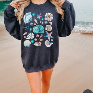 May include: Dark heather sweatshirt featuring a collection of colorful seashells and starfish. The design includes various shell shapes and sizes in shades of blue, pink, and white, creating a coastal theme. The sweatshirt is a comfortable, casual style.