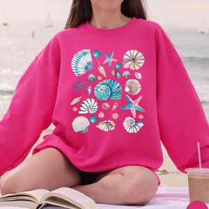 May include: A bright pink sweatshirt featuring a collection of watercolor seashells and starfish in shades of blue, white, and tan. The design includes various shell shapes and sizes, creating a coastal theme.