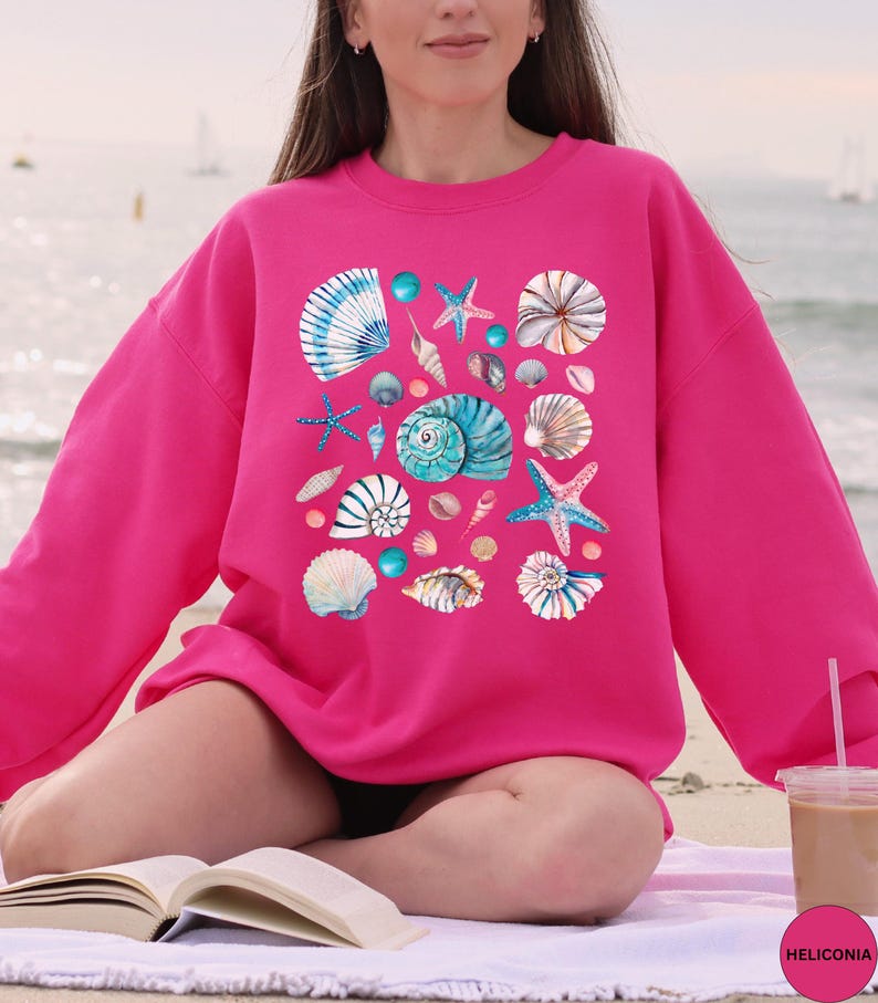 May include: A bright pink sweatshirt featuring a collection of watercolor seashells and starfish in shades of blue, white, and tan. The design includes various shell shapes and sizes, creating a coastal theme.