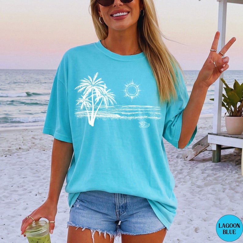 May include: A lagoon blue short-sleeved t-shirt featuring a white graphic print of palm trees, a sun, and ocean waves on the front. The shirt has a crew neckline and a relaxed fit, showcasing a coastal scene with a clear sky and water.