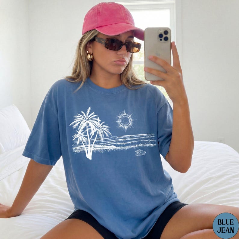 May include: A blue short-sleeve t-shirt featuring a white line art graphic of palm trees, a sun, and an ocean shoreline. The shirt is paired with a pink baseball cap and dark-tinted sunglasses. The text BLUE JEAN is visible in the bottom corner.