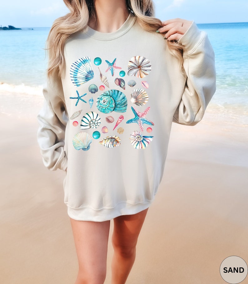 May include: A sand-colored sweatshirt featuring a watercolor design of various seashells, starfish, and beads in shades of blue, pink, and white. The design is centered on the front of the long-sleeved top. The word "SAND" is printed in a circle at the bottom right.