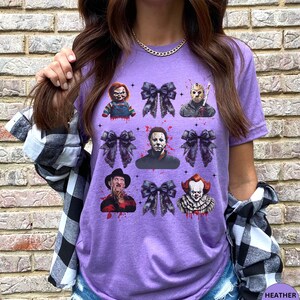 May include: Purple t-shirt featuring a collage of horror movie characters and black bows. The design includes images of Chucky, Jason Voorhees, Michael Myers, Freddy Krueger, and Pennywise, with blood splatter accents.