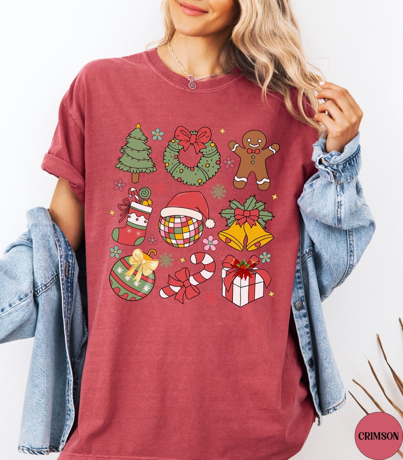 May include: A crimson short-sleeve t-shirt featuring a grid of colourful Christmas-themed illustrations including a tree, wreath, gingerbread person, stocking, disco ball with a Santa hat, bells, ornament, candy cane, and a gift box.