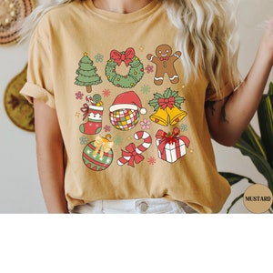 May include: A mustard yellow t-shirt featuring a graphic print of various Christmas icons including a tree, wreath, gingerbread person, stocking, disco ball with a Santa hat, bells, ornament, candy cane, and a gift box with a red bow.