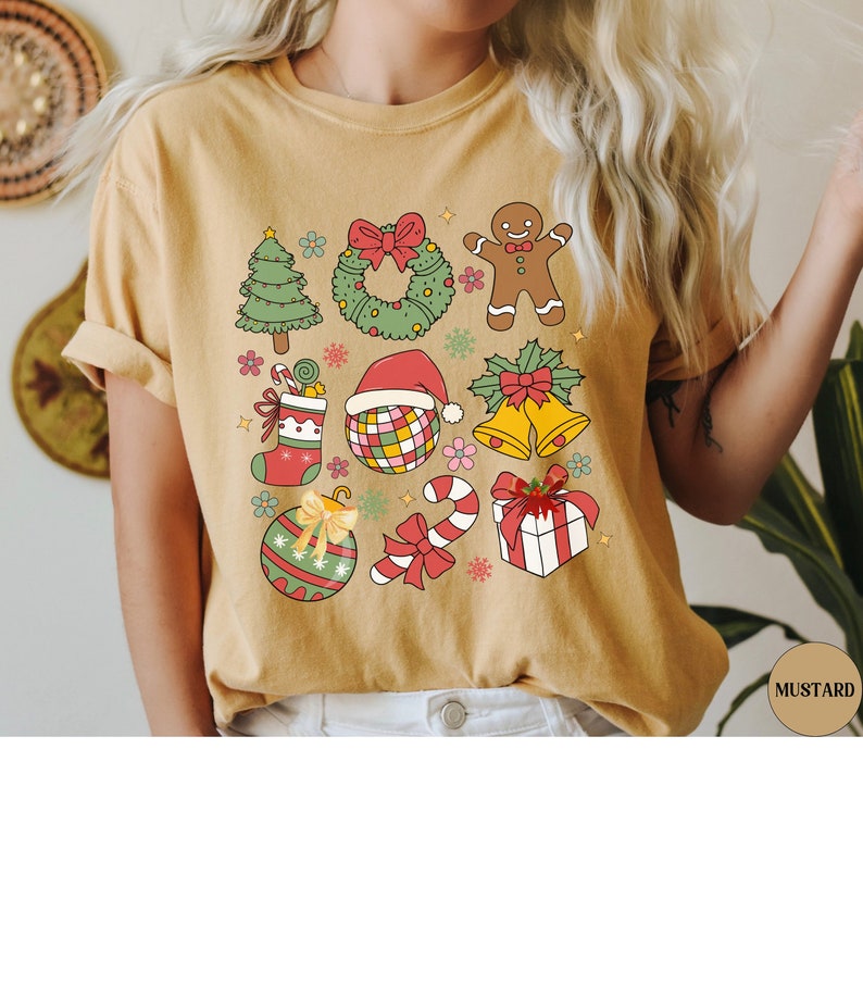 May include: A mustard yellow t-shirt featuring a graphic print of various Christmas icons including a tree, wreath, gingerbread person, stocking, disco ball with a Santa hat, bells, ornament, candy cane, and a gift box with a red bow.