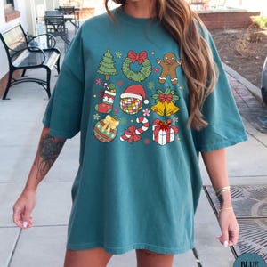 May include: A teal short-sleeve t-shirt featuring a grid of nine colourful Christmas-themed illustrations, including a pine tree, a wreath, a gingerbread man, a stocking, a disco ball with a Santa hat, bells, an ornament, a candy cane, and a gift.
