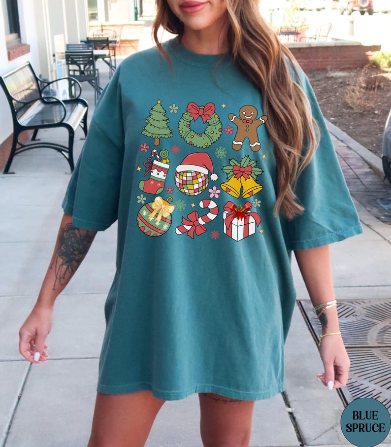 May include: A teal short-sleeve t-shirt featuring a grid of nine colourful Christmas-themed illustrations, including a pine tree, a wreath, a gingerbread man, a stocking, a disco ball with a Santa hat, bells, an ornament, a candy cane, and a gift.
