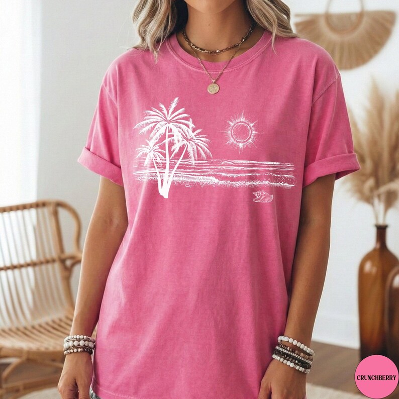 May include: A bright pink short-sleeved t-shirt featuring a white graphic print of palm trees, a sun, and ocean waves on the front. The shirt has a crew neckline and a relaxed fit, showcasing a beach-themed illustration centred on the chest area.