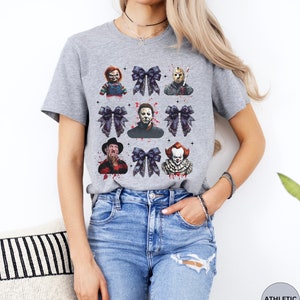May include: Athletic heather gray t-shirt featuring a grid of horror movie characters and black bows. The characters include Chucky, Jason Voorhees, Michael Myers, Freddy Krueger, and Pennywise. The shirt is made of a soft, comfortable material.