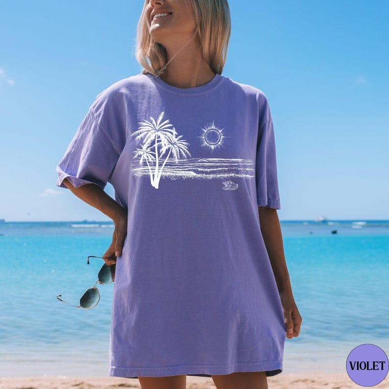 May include: A violet short-sleeved t-shirt featuring a white graphic print of palm trees, a sun, and ocean waves on the front. The shirt has a crew neckline and a relaxed fit, with a small circular violet icon in the corner labelled VIOLET.