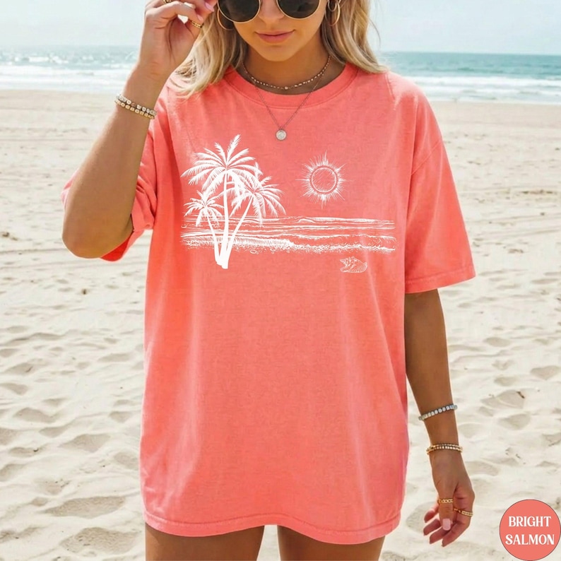 May include: A coral-coloured short-sleeve t-shirt featuring a white graphic print on the chest. The design depicts a beach scene with palm trees, a sun, and ocean waves. The shirt has a crew neckline and a relaxed fit, made of a solid fabric.