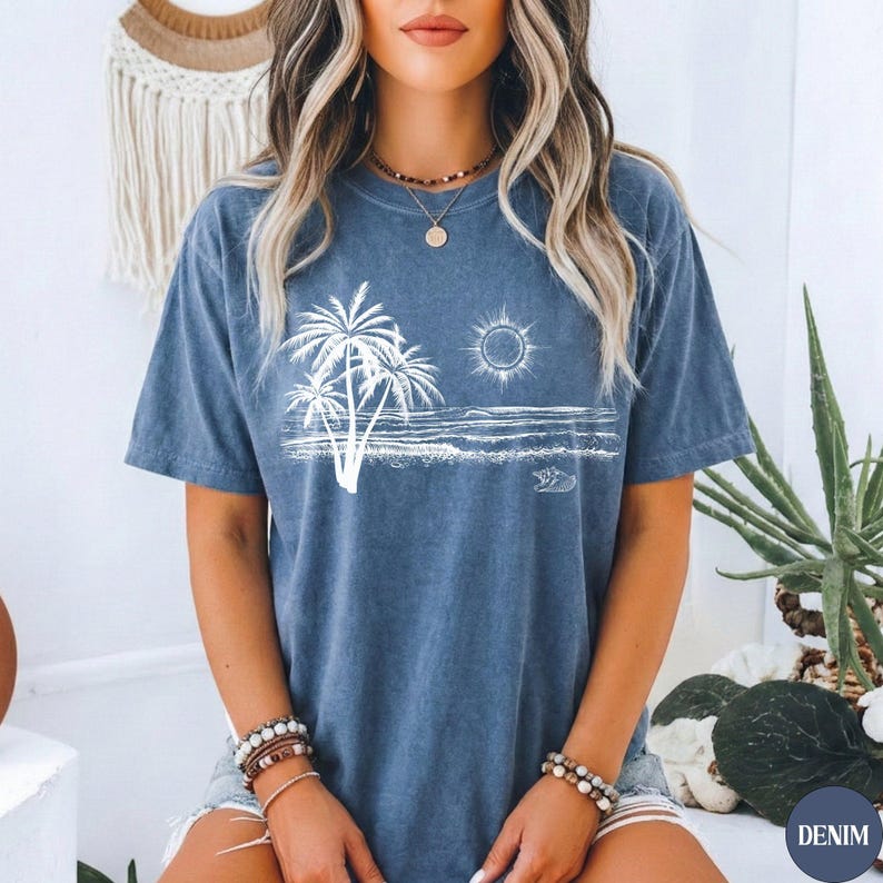 May include: A blue short-sleeve t-shirt featuring a white graphic print of palm trees, a sun, and ocean waves. The shirt has a crew neckline and a washed-out, vintage-inspired texture. The design is centred on the chest area of the garment.