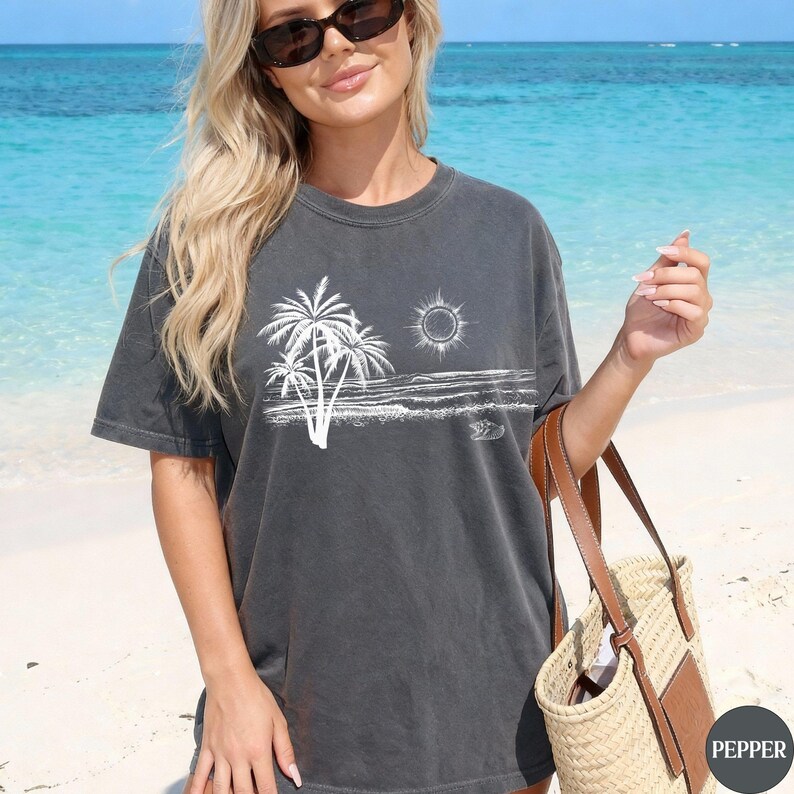 May include: A charcoal grey short-sleeve t-shirt featuring a white line art graphic of palm trees, a sun, and ocean waves. The shirt has a crew neckline and a relaxed fit, showcasing a beach-themed illustration printed on the front chest area.