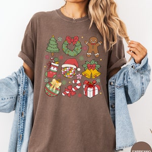 May include: A brown t-shirt featuring a grid of colourful Christmas-themed illustrations including a tree, wreath, gingerbread man, stocking, disco ball with a Santa hat, bells, ornament, candy cane, and a gift box with a red bow.