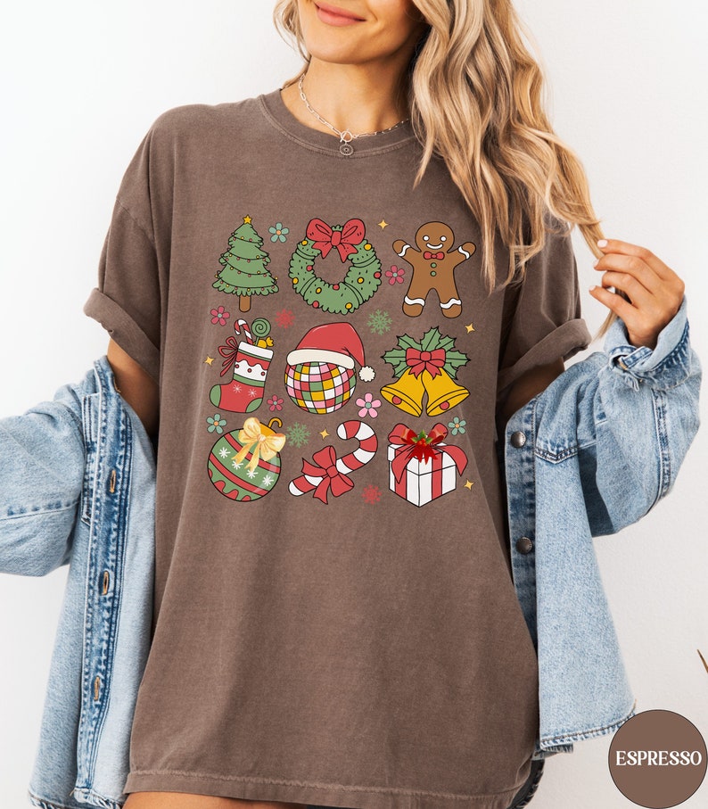 May include: A brown t-shirt featuring a grid of colourful Christmas-themed illustrations including a tree, wreath, gingerbread man, stocking, disco ball with a Santa hat, bells, ornament, candy cane, and a gift box with a red bow.