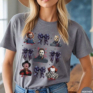 May include: A gray t-shirt featuring a design with horror movie characters and dark blue bows. The characters include Chucky, Jason Voorhees, Michael Myers, Freddy Krueger, and Pennywise. The text "HEATHER STORM" is visible.