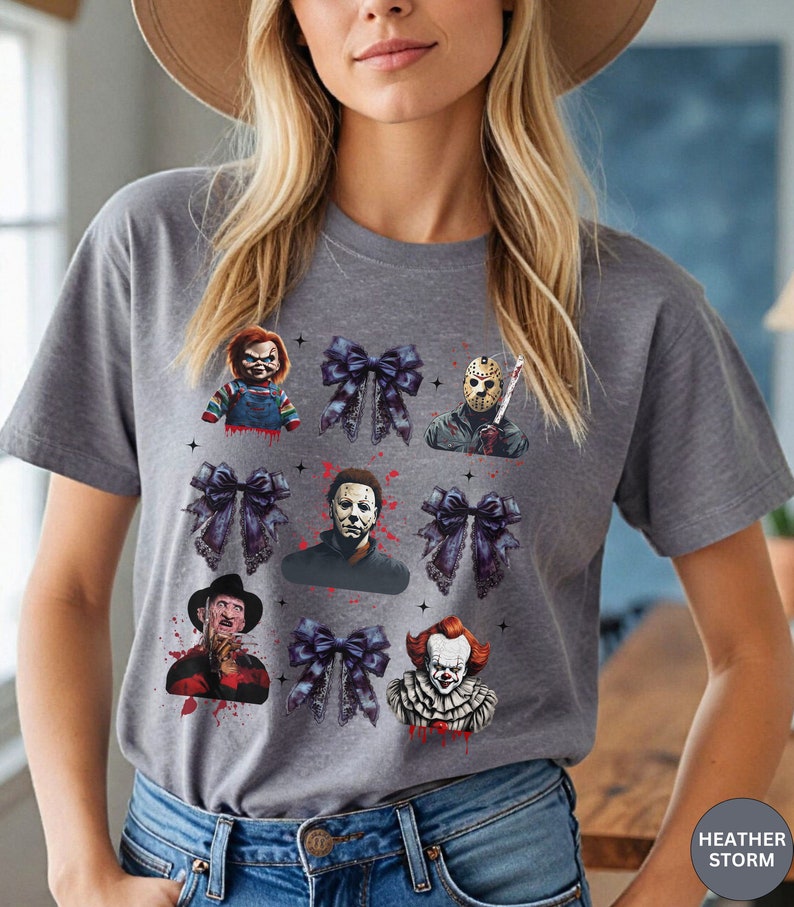 May include: A gray t-shirt featuring a design with horror movie characters and dark blue bows. The characters include Chucky, Jason Voorhees, Michael Myers, Freddy Krueger, and Pennywise. The text "HEATHER STORM" is visible.