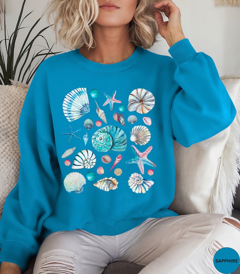 May include: A turquoise blue sweatshirt with a colorful seashell and starfish design. The design features a variety of seashells in different shapes and sizes, as well as starfish.