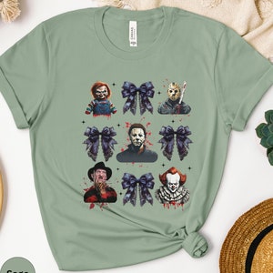 May include: Sage green t-shirt featuring a grid design with horror movie characters and dark blue bows. The characters include Chucky, Jason Voorhees, Michael Myers, Freddy Krueger, and Pennywise. The word "Sage" is printed on the bottom left.