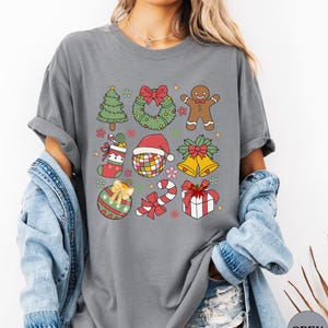 May include: A charcoal grey t-shirt featuring a grid of colourful Christmas-themed illustrations including a tree, wreath, gingerbread person, stocking, disco ball with a Santa hat, bells, ornament, candy cane, and a gift box with a red bow.
