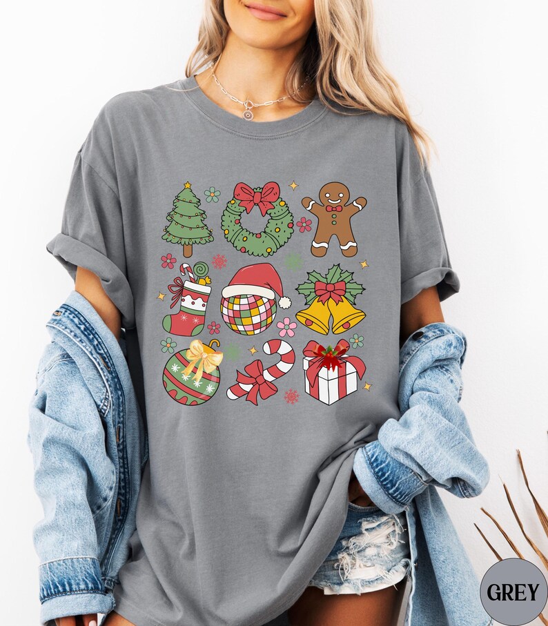 May include: A charcoal grey t-shirt featuring a grid of colourful Christmas-themed illustrations including a tree, wreath, gingerbread person, stocking, disco ball with a Santa hat, bells, ornament, candy cane, and a gift box with a red bow.