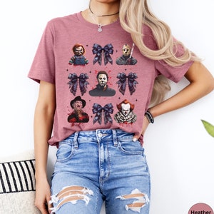 May include: Heather mauve t-shirt featuring a grid of horror movie characters and dark blue bows. The characters include Chucky, Jason Voorhees, Michael Myers, Freddy Krueger, and Pennywise. The shirt has a crew neck and short sleeves.