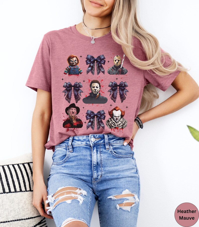 May include: Heather mauve t-shirt featuring a grid of horror movie characters and dark blue bows. The characters include Chucky, Jason Voorhees, Michael Myers, Freddy Krueger, and Pennywise. The shirt has a crew neck and short sleeves.