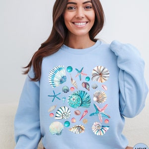 May include: Light blue sweatshirt featuring a watercolor design of various seashells, starfish, and teal-colored beads. The design includes a variety of shell shapes and sizes, with blue, white, and pink accents. The sweatshirt has a crew neck and long sleeves.