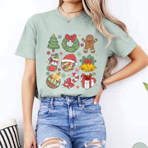 May include: A sage green short-sleeve t-shirt featuring a graphic print of various Christmas icons including a tree, wreath, gingerbread person, stocking, disco ball with a Santa hat, bells, ornament, candy cane, and a gift box with a red bow.