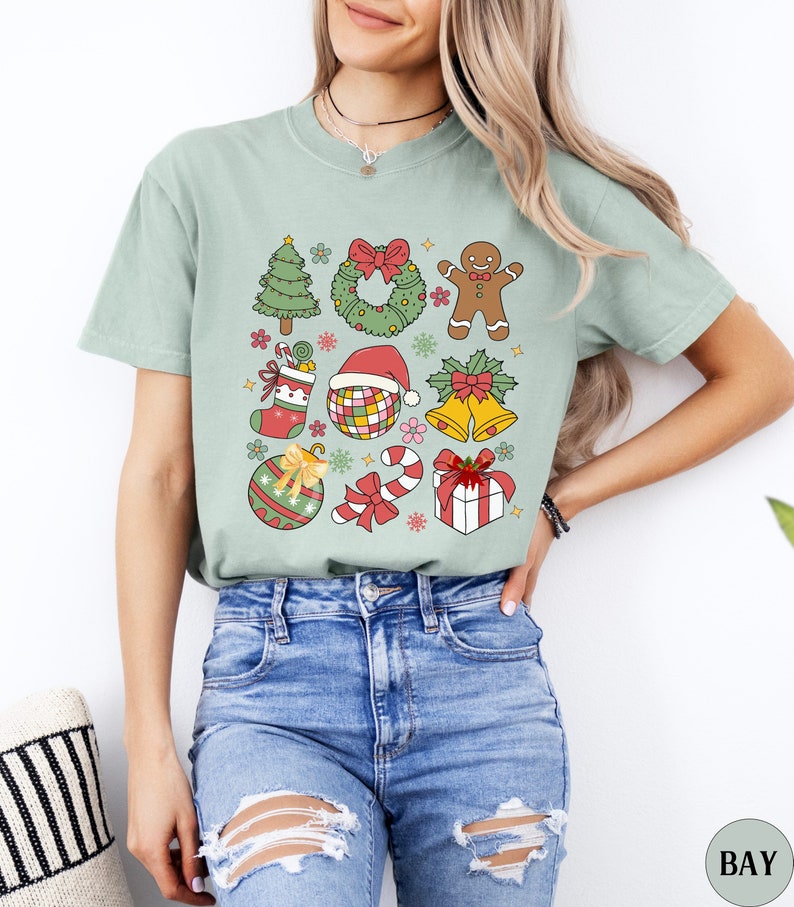 May include: A sage green short-sleeve t-shirt featuring a graphic print of various Christmas icons including a tree, wreath, gingerbread person, stocking, disco ball with a Santa hat, bells, ornament, candy cane, and a gift box with a red bow.