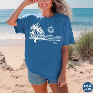 May include: A blue short-sleeved t-shirt featuring a white graphic print of a beach scene with palm trees, a sun, ocean waves, and a small seashell. The design is centred on the chest area of the garment, which has a crew neckline and loose fit.