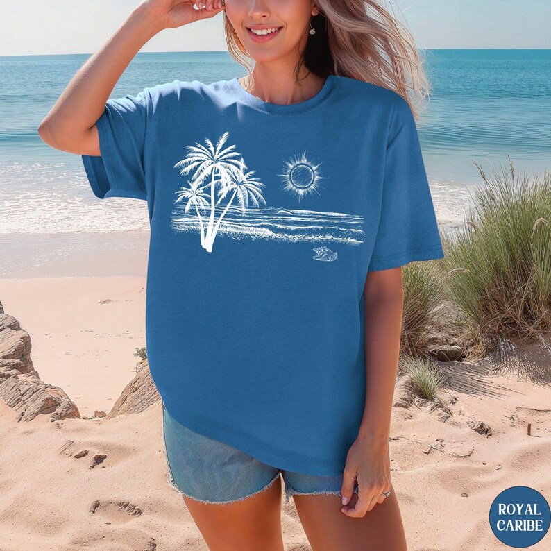 May include: A blue short-sleeved t-shirt featuring a white graphic print of a beach scene with palm trees, a sun, ocean waves, and a small seashell. The design is centred on the chest area of the garment, which has a crew neckline and loose fit.