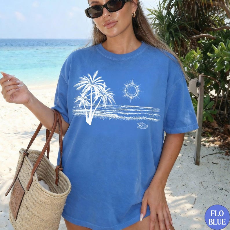 May include: A blue short-sleeve t-shirt featuring a white graphic print of palm trees, a sun, and ocean waves on the front. The shirt has a crew neckline and a relaxed fit. A woven straw tote bag with brown leather handles is held at the side.